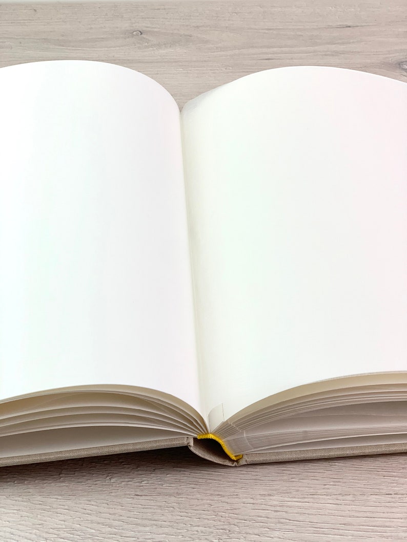 May include: A blank, white book with a linen cover and a yellow ribbon bookmark. The book is open and lying flat on a light wood surface.