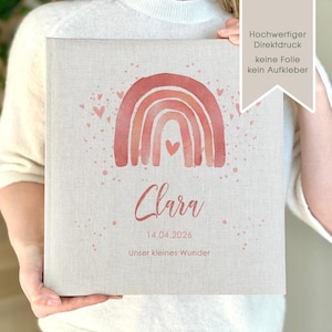 May include: A personalized photo album with a watercolor rainbow design in shades of pink and orange. The album has the name "Clara" and the date "14.04.2026" printed on it. The text "Unser kleines Wunder" is also printed on the album.