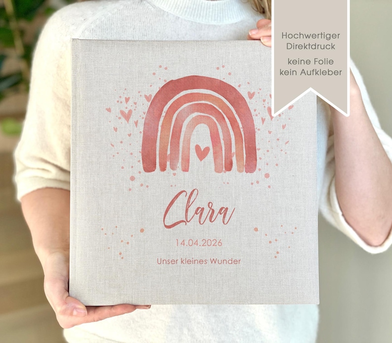 May include: A personalized photo album with a watercolor rainbow design in shades of pink and orange. The album has the name "Clara" and the date "14.04.2026" printed on it. The text "Unser kleines Wunder" is also printed on the album.