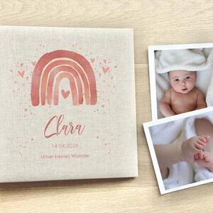 May include: A pink and white linen baby photo album with a watercolor rainbow and hearts. The album has the name "Clara" and the date "14.04.2026" printed on it. The text "Unser kleines Wunder" is also printed on the album.