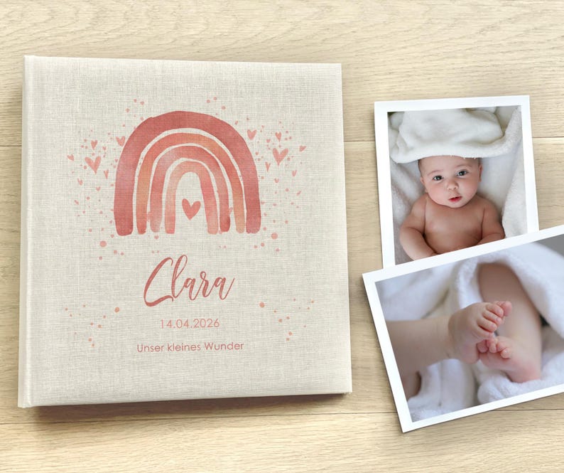 May include: A pink and white linen baby photo album with a watercolor rainbow and hearts. The album has the name "Clara" and the date "14.04.2026" printed on it. The text "Unser kleines Wunder" is also printed on the album.
