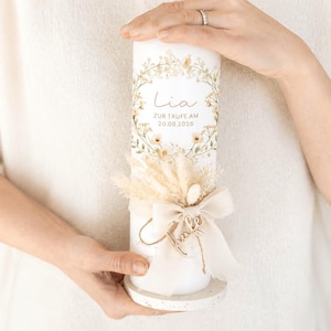 May include: A white cylindrical candle featuring a floral wreath design, the name Lia, and the text ZUR TAUFE AM 20.08.2025. The candle is decorated with a beige ribbon bow, dried grasses, and a small wooden name tag, resting on a speckled base.