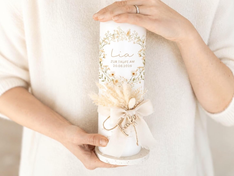 May include: A white cylindrical candle featuring a floral wreath design, the name Lia, and the text ZUR TAUFE AM 20.08.2025. The candle is decorated with a beige ribbon bow, dried grasses, and a small wooden name tag, resting on a speckled base.