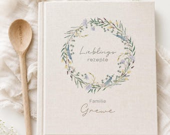 May include: A square, beige fabric-covered recipe book featuring a watercolor floral wreath illustration surrounding the text "Lieblings rezepte" and "Familie Grewe" below, accompanied by a light-colored wooden spoon with "MARLEY & MORE" engraved on the handle.