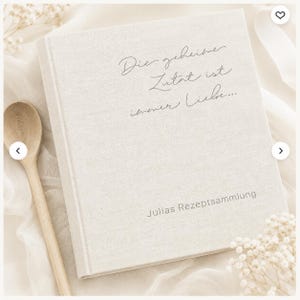May include: A square, light beige fabric-covered recipe book with the German text "Die geheime Zutat ist immer Liebe..." and "Julias Rezeptsammlung" printed on the front cover, accompanied by a wooden spoon placed to the left of the book.