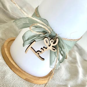 May include: A white candle is adorned with a sage green ribbon and twine, with a wooden cutout that reads "Taufe". The candle sits on a heart-shaped wooden base. The overall aesthetic is soft and natural.