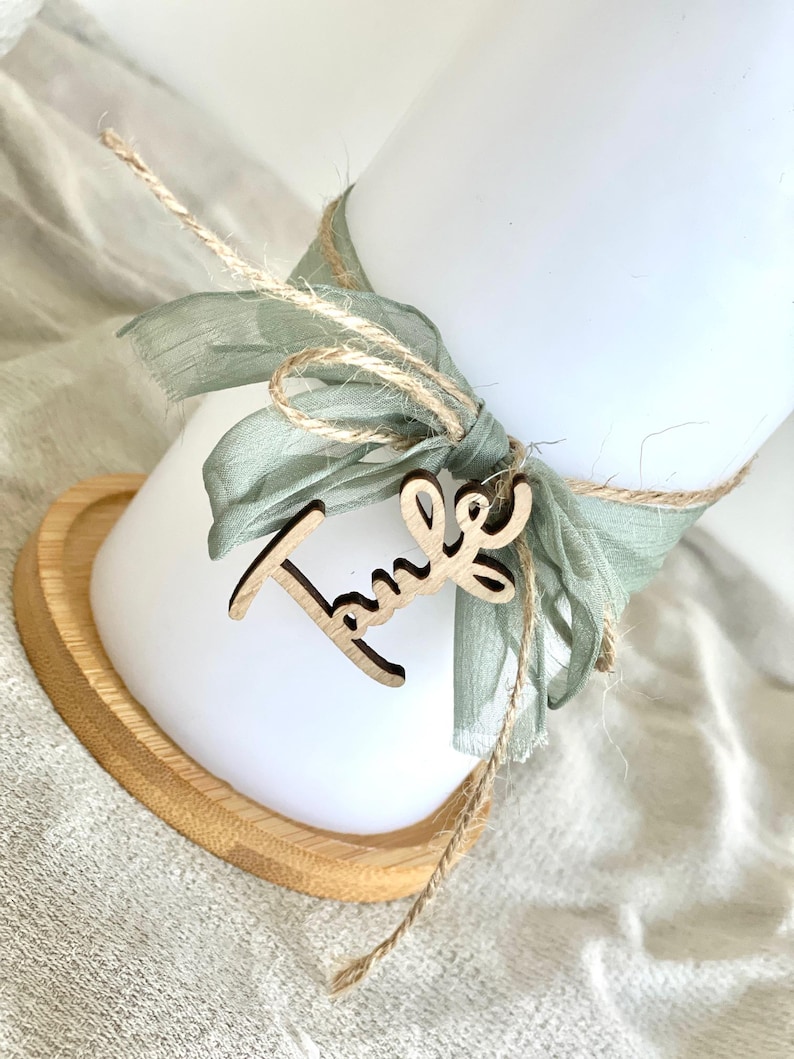 May include: A white candle is adorned with a sage green ribbon and twine, with a wooden cutout that reads "Taufe". The candle sits on a heart-shaped wooden base. The overall aesthetic is soft and natural.