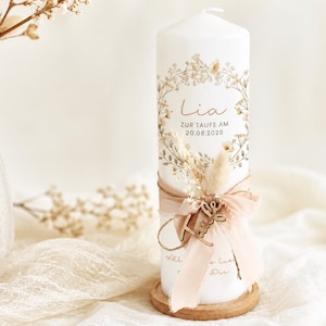 May include: A white pillar candle featuring a floral wreath graphic, the name Lia, and the text ZUR TAUFE AM 20.08.2025. The candle is decorated with a beige ribbon, dried grasses, and a small wooden charm, resting on a circular wooden base.
