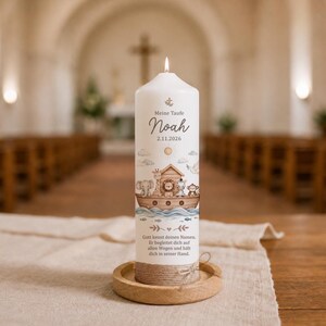 Könnte beinhalten: Eine weiße zylindrische Kerze mit brennendem Docht, bedruckt mit einer Arche Noah Illustration, Tieren, dem Text "Meine Taufe", "Noah", "2.11.2026" sowie einem religiösen Spruch. Der Fuß ist mit Kordel umwickelt und steht auf einem Holztablett.