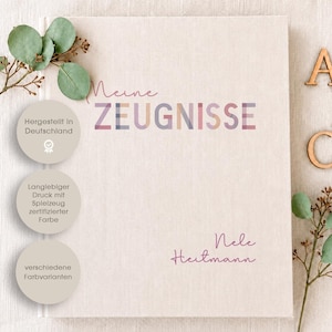 Könnte beinhalten: Ein hellbeiges, stoffbezogenes Buch mit dem aufgedruckten Text Meine ZEUGNISSE und dem Namen Nele Heitmann. Drei kreisförmige Abzeichen listen deutsche Produktionsdetails auf, und hölzerne Buchstaben A, B und C liegen neben grünen Eukalyptuszweigen.