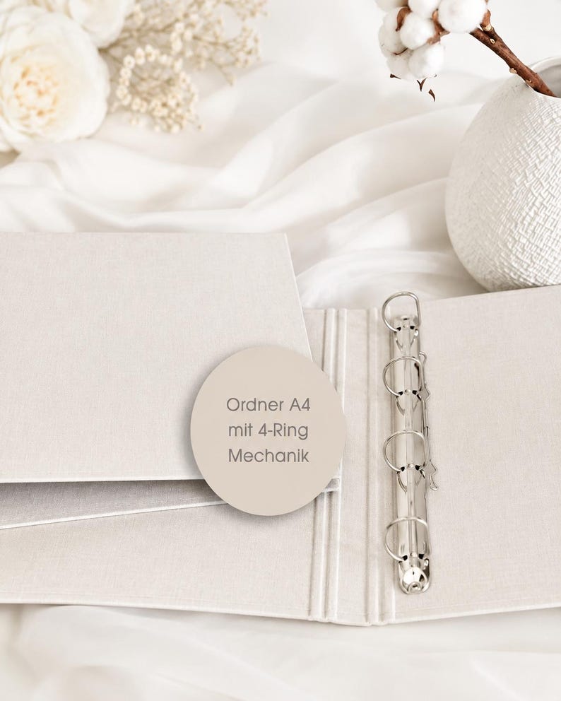 May include: A light beige A4 binder featuring a silver-colored 4-ring metal mechanism. A circular sticker is placed on the cover with the text Ordner A4 mit 4-Ring Mechanik printed in a dark font, indicating the product type and its internal hardware.
