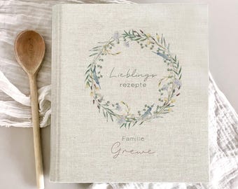 May include: A linen-covered cookbook with the words "Lieblingsrezepte Familie Grewe" in script. A floral wreath design in green, blue, and purple encircles the title. A wooden spoon with the text "MARLEY COOK" is next to the book.