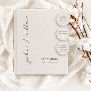 Könnte beinhalten: Ein beigefarbenes Stammbuch mit Leinenbezug, das den vertikalen Schriftzug "julia & niklas" sowie den gedruckten Text "STAMMBUCH FAMILIE KAUER" trägt. Drei runde Aufkleber enthalten Informationen zu Druckqualität, Herkunft und Material.