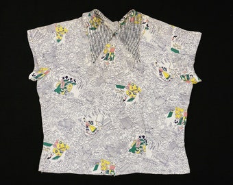 Pode incluir: Uma blusa branca de manga curta com um design estampado com detalhes em verde, amarelo e rosa. A blusa tem um colar com listras pretas e brancas.
