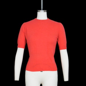 May include: A coral colored short-sleeved sweater on a white mannequin.
