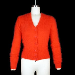 Puede incluir: Un cárdigan naranja brillante con cuello en V y mangas largas. El tejido de punto difuso tiene una parte delantera abotonada y se muestra en un maniquí sobre un fondo negro. Este suéter de estilo vintage es una adición acogedora a cualquier guardarropa.