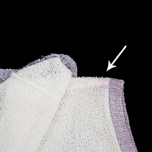 May include: Close-up of a white and purple knitted fabric with a frayed edge. The fabric is folded over, and the arrow points to the edge of the fabric.
