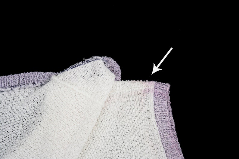 May include: Close-up of a white and purple knitted fabric with a frayed edge. The fabric is folded over, and the arrow points to the edge of the fabric.