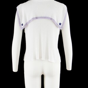 May include: White sleeveless sweater with a lavender trim and blue star accents. The sweater has a sailor collar.