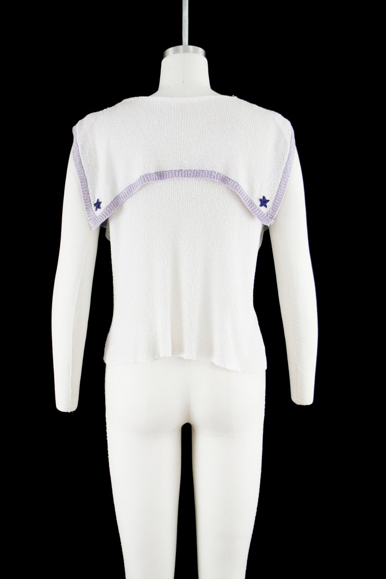 May include: White sleeveless sweater with a lavender trim and blue star accents. The sweater has a sailor collar.
