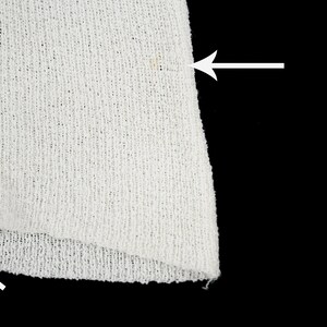 May include: Close-up of a white knitted fabric with a small brown stain. The fabric has a loose weave and a slightly frayed edge.