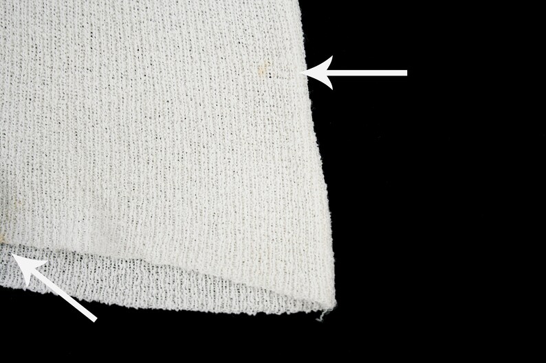 May include: Close-up of a white knitted fabric with a small brown stain. The fabric has a loose weave and a slightly frayed edge.