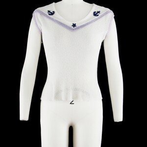 May include: White sleeveless sweater with a purple and blue trim around the neckline. The neckline has a star and two anchors in the center.