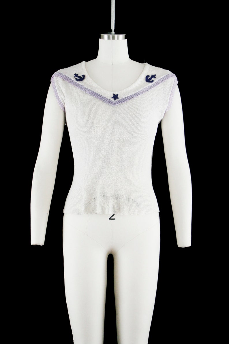 May include: White sleeveless sweater with a purple and blue trim around the neckline. The neckline has a star and two anchors in the center.