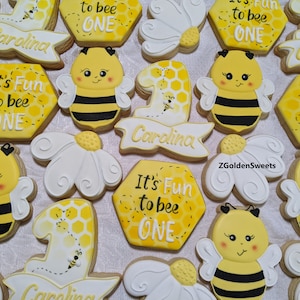 May include: A set of decorated sugar cookies featuring a bee theme. The cookies are decorated with yellow and black stripes, white wings, and a honeycomb pattern. The cookies also feature the text "It's Fun to bee One" and "Carolina".