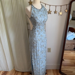 May include: A pale blue halter-neck evening gown with detailed floral embroidery and silver beading. The dress has a fitted bodice, a flowing skirt, and a high slit. The dress is displayed on a mannequin.