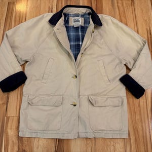 May include: A tan jacket with a navy blue collar and cuffs. The jacket has two front pockets and a blue plaid lining visible at the collar. The jacket is buttoned up and appears to be made of a lightweight material.