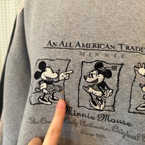 May include: Gray sweatshirt featuring embroidered Minnie Mouse designs. The text "An All American Tradition" and "Minnie Mouse" are above and below the images. The sweatshirt also includes the text "The One Only Authentic, Original Minnie Since 1928".