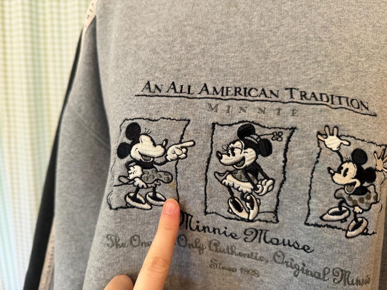 May include: Gray sweatshirt featuring embroidered Minnie Mouse designs. The text "An All American Tradition" and "Minnie Mouse" are above and below the images. The sweatshirt also includes the text "The One Only Authentic, Original Minnie Since 1928".