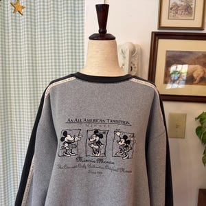 May include: A gray sweatshirt with black trim and sleeves. The front features a graphic of Minnie Mouse and the text "An All American Tradition Minnie Mouse". The sleeves have a black stripe with white text.