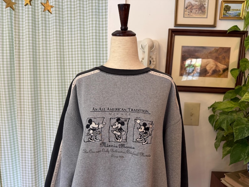 May include: A gray sweatshirt with black trim and sleeves. The front features a graphic of Minnie Mouse and the text "An All American Tradition Minnie Mouse". The sleeves have a black stripe with white text.