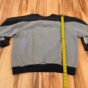 May include: A gray and black sweatshirt with a crew neck. The top portion of the sweatshirt is black, while the body is gray. The cuffs and bottom hem are black. A yellow measuring tape is placed vertically along the side of the sweatshirt.