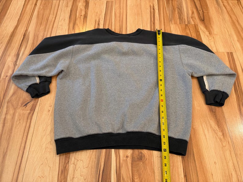 May include: A gray and black sweatshirt with a crew neck. The top portion of the sweatshirt is black, while the body is gray. The cuffs and bottom hem are black. A yellow measuring tape is placed vertically along the side of the sweatshirt.