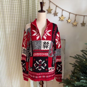 Puede incluir: Un suéter estampado en rojo, negro y blanco con cuello con cremallera. El suéter presenta diseños de copos de nieve y geométricos. El suéter se exhibe en un maniquí. El diseño del suéter recuerda un tema festivo.