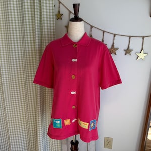 Op de afbeelding: Een felroze shirt met korte mouwen, kraag en knoopsluiting. Decoratieve patches met vissen, zonnen en andere ontwerpen sieren de zoom. De knopen zijn bruin en wit.