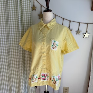 Op de afbeelding: Een gele shirt met korte mouwen met geborduurde fruitontwerpen. De shirt heeft een zak met ananasborduurwerk en een zoom met ananas-, watermeloen- en kersmotieven. De shirt wordt op een mannequin getoond.