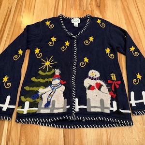 May include: Navy blue button-up cardigan sweater with a festive Christmas theme. The sweater features embroidered yellow stars, a Christmas tree, and snowmen. White trim accents the collar, cuffs, and hem, creating a cozy winter look.