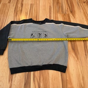 May include: Gray sweatshirt with black sleeves and trim. The sweatshirt features a graphic of Mickey Mouse and the text "An All American Tradition". The length of the sweatshirt is being measured with a tape measure.