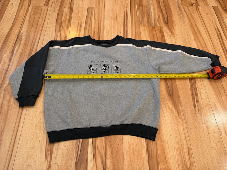 May include: Gray sweatshirt with black sleeves and trim. The sweatshirt features a graphic of Mickey Mouse and the text "An All American Tradition". The length of the sweatshirt is being measured with a tape measure.