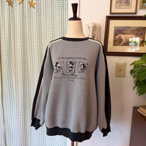 May include: Gray sweatshirt with black sleeves and trim. The front features a graphic of Minnie Mouse and the text "An All American Tradition." The sweatshirt is displayed on a mannequin.