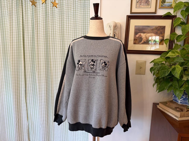 May include: Gray sweatshirt with black sleeves and trim. The front features a graphic of Minnie Mouse and the text "An All American Tradition." The sweatshirt is displayed on a mannequin.