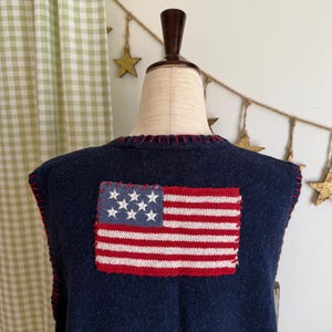 Puede incluir: Un chaleco azul marino con un parche de la bandera estadounidense cosido en la espalda. La bandera presenta estrellas blancas sobre un campo azul y rayas rojas y blancas. El chaleco tiene costuras rojas en los bordes.