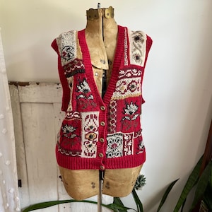Op de afbeelding: Een rode vest met knopen en een V-hals, weergegeven op een vintage paspop. Het vest heeft een patchwork-ontwerp met verschillende patronen in crème, zwart en rood. Het vest heeft een geribbelde zoom en mouwopeningen.