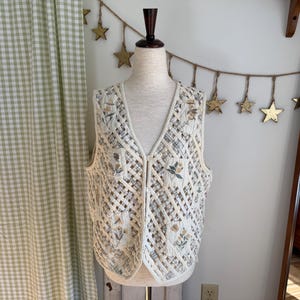 May include: Cream-colored quilted vest with a lattice design and floral embroidery. The vest has a V-neck and a button-up front. The background includes a green and white checkered curtain and a gold star garland.