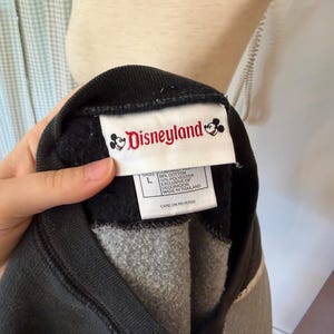 May include: A black and gray Disneyland sweatshirt with a white tag featuring the Disneyland logo and text. The sweatshirt is a size large and appears to be made of a soft, comfortable material. The tag also includes information about the materials used.