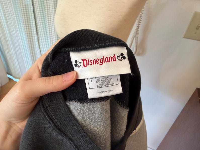 May include: A black and gray Disneyland sweatshirt with a white tag featuring the Disneyland logo and text. The sweatshirt is a size large and appears to be made of a soft, comfortable material. The tag also includes information about the materials used.